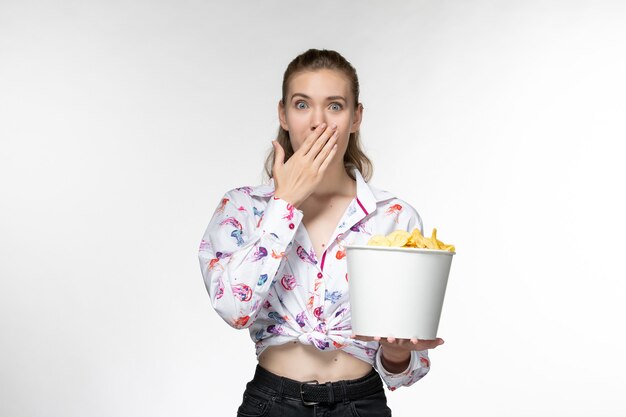 Vue de face jeune belle femme tenant des cips de pomme de terre regarder un film sur une surface blanche légère