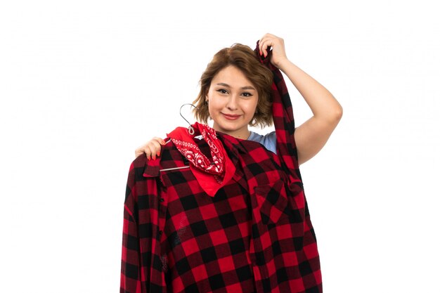 Une vue de face jeune belle fille en t-shirt bleu et jean bleu tenant une chemise à carreaux rouge-noir souriant sur le blanc