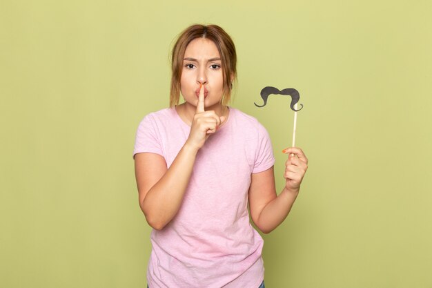 Une vue de face jeune belle fille en t-shirt rose jeans bleu posant avec une fausse moustache montrant signe de silence sur le vert