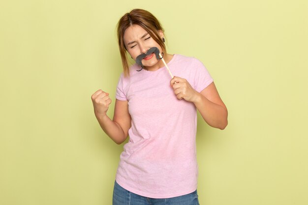 Une vue de face jeune belle fille en t-shirt rose jeans bleu posant avec une fausse moustache sur vert