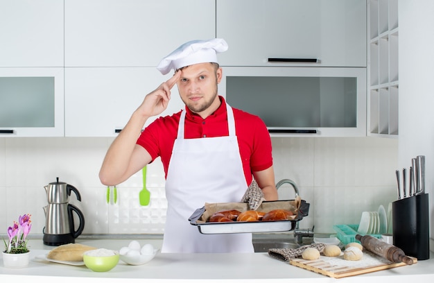 Vue de face d'un jeune chef masculin portant un support tenant des pâtisseries fraîchement préparées et se sentant fier dans la cuisine blanche