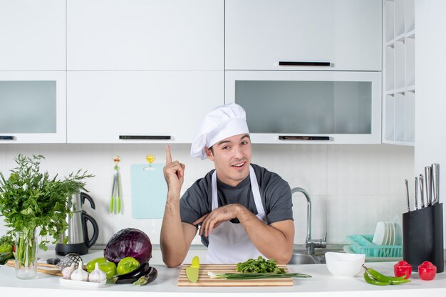 Vue de face jeune chef en uniforme de coupe de légumes verts sur une planche à découper