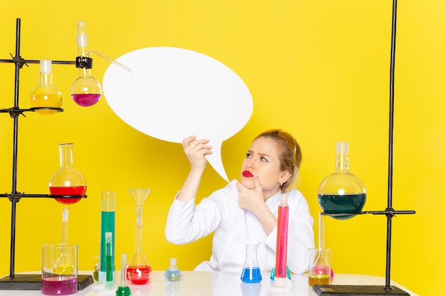 Photo gratuite vue de face jeune chimiste assis en costume blanc avec différentes solutions tenant une pancarte blanche sur le travail de processus scientifique de chimie de l'espace jaune