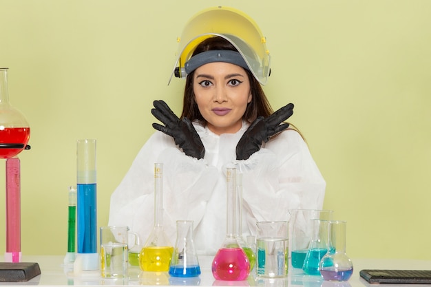 Vue de face jeune chimiste en combinaison de protection spéciale assis en face de la table avec des solutions sur le mur vert travail chimie laboratoire chimique science féminine