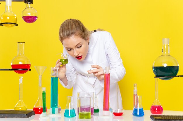 Vue de face jeune chimiste en costume blanc en face de la table avec des solutions ed sur l'expérience scientifique de chimie de l'espace jaune