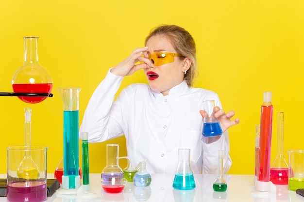 Vue de face jeune chimiste en costume blanc en face de la table avec des solutions ed holdign son nez sur la science de la chimie de l'espace jaune