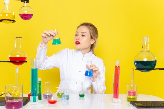 Vue de face jeune chimiste en costume blanc en face de la table avec des solutions ed tenant un sur l'expérience scientifique de chimie de l'espace jaune