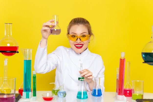 Vue de face jeune chimiste en costume blanc en face de la table avec des solutions ed travaillant avec eux sur la chimie de l'espace jaune