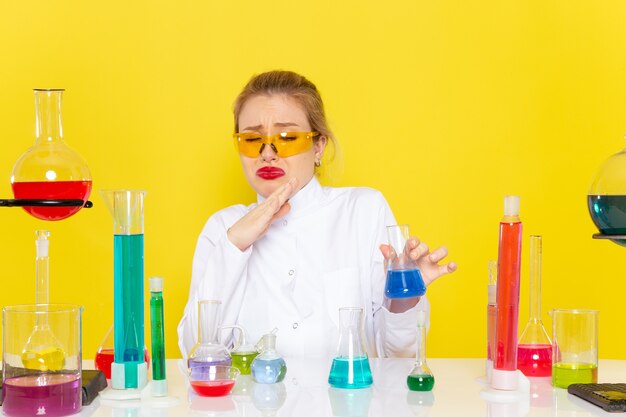 Vue de face jeune chimiste en costume blanc en face de la table avec des solutions ed travaillant avec eux sur le travail de chimie de l'espace jaune