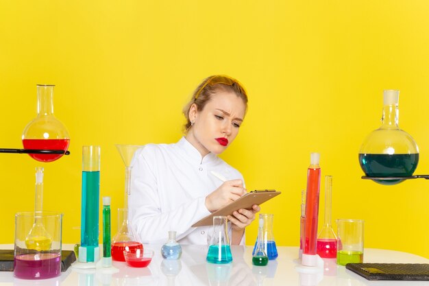 Vue de face jeune chimiste en costume blanc avec des solutions ed travaillant avec eux et assis à écrire sur la chimie de l'espace jaune