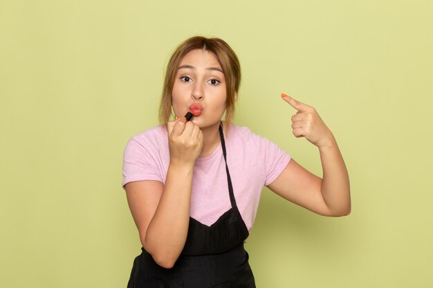 Une vue de face jeune coiffeuse en t-shirt rose et cape noire faisant du maquillage sur le vert