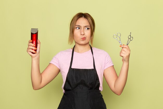 Une vue de face jeune coiffeuse en t-shirt rose et cape noire tenant une machine à cheveux et des ciseaux sur vert