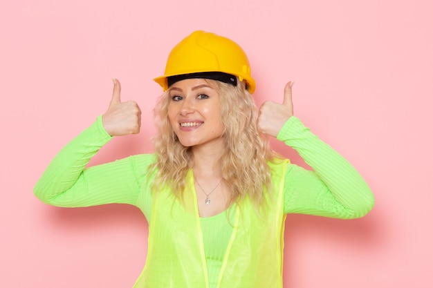 Photo gratuite vue de face jeune constructeur féminin en costume de construction vert casque jaune posant juste avec le sourire sur le travail de construction de l'architecture de l'espace rose