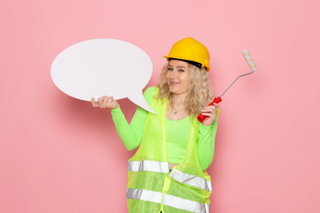 Vue de face jeune constructeur féminin en costume de construction verte casque tenant une pancarte blanche et souriant juste sur les travaux de construction de l'architecture de l'espace rose