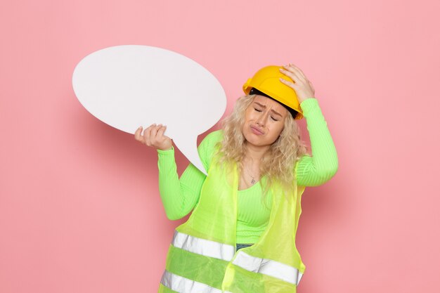 Vue de face jeune constructeur féminin en costume de construction verte casque tenant panneau blanc déprimé sur l'espace rose