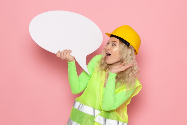 Vue de face jeune constructeur féminin en costume de construction verte casque tenant panneau blanc surpris sur l'espace rose