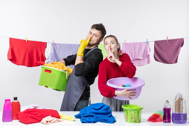 Vue de face d'un jeune couple heureux faisant un signe de silence tenant des trucs à laver sur un mur blanc