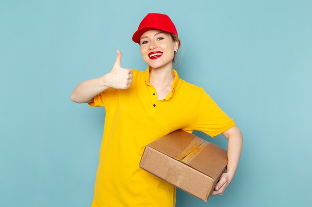 Vue de face jeune courrier féminin en chemise jaune et cape rouge holding package souriant sur le travail de l'espace bleu