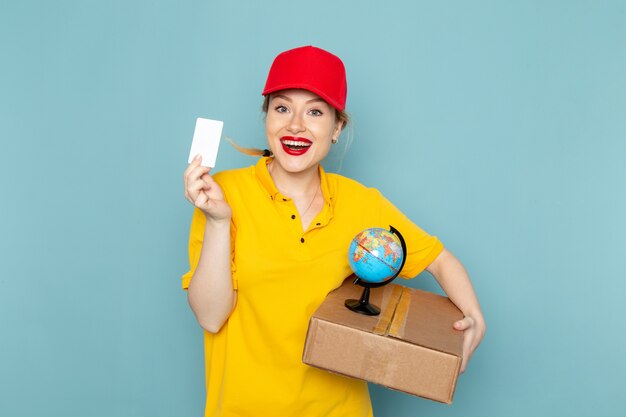 Vue de face jeune courrier féminin en chemise jaune et cape rouge tenant petite carte globe et paquet souriant sur l'espace bleu