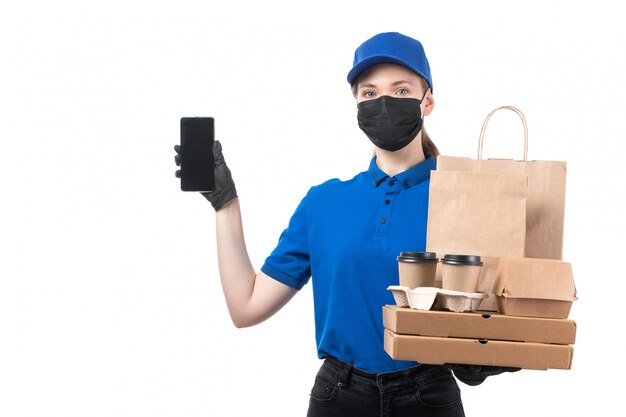 Une vue de face jeune courrier en uniforme bleu gants noirs et masque noir tenant des colis de livraison de nourriture et smartphone