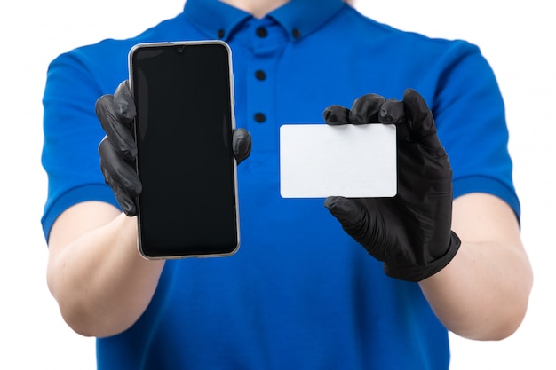 Une vue de face jeune courrier en uniforme bleu gants noirs et masque noir tenant téléphone et carte blanche