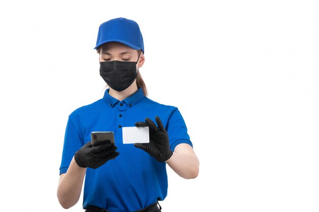 Une vue de face jeune courrier en uniforme bleu gants noirs et masque noir tenant téléphone et carte blanche