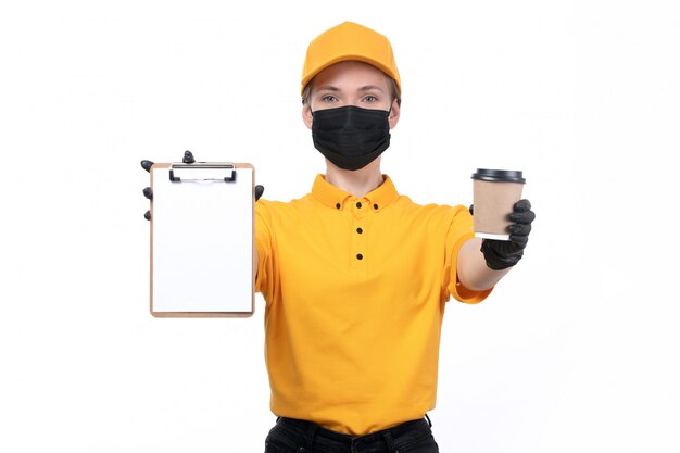 Une Vue De Face Jeune Courrier En Uniforme Jaune Gants Noirs Et Masque Noir Tenant Le Bloc-notes Avec Tasse à Café