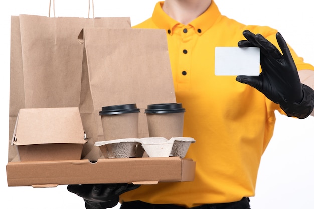 Une vue de face jeune courrier en uniforme jaune gants noirs et masque noir tenant des boîtes à pizza et carte blanche offrant
