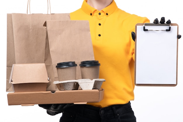 Photo gratuite une vue de face jeune courrier en uniforme jaune gants noirs et masque noir tenant des paquets de nourriture et des tasses à café