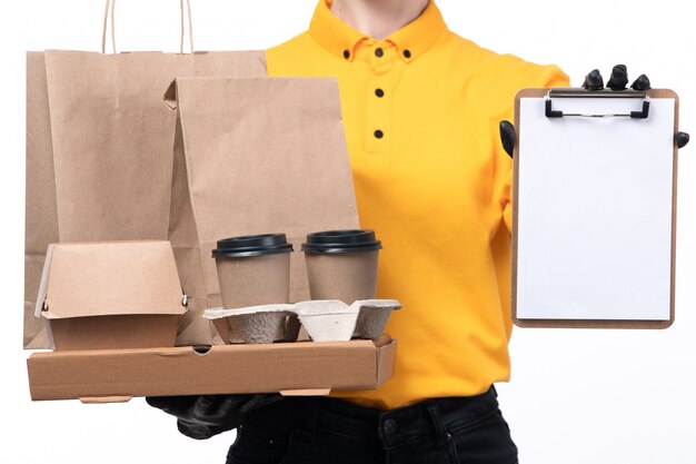 Une vue de face jeune courrier en uniforme jaune gants noirs et masque noir tenant des paquets de nourriture et des tasses à café