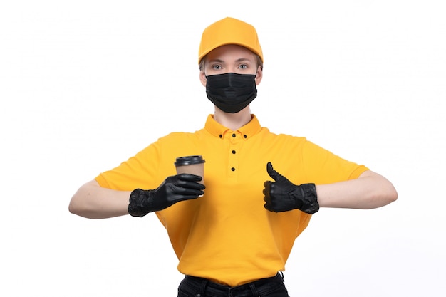 Une vue de face jeune courrier en uniforme jaune gants noirs et masque noir tenant une tasse de café montrant comme signe