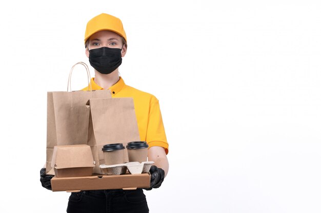 Une vue de face jeune courrier en uniforme jaune gants noirs et masque noir tenant des tasses à café et des paquets de livraison de commande