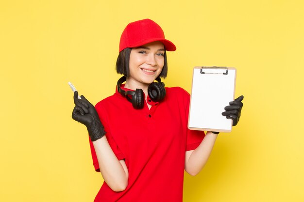 Une vue de face jeune courrier en uniforme rouge gants noirs et bonnet rouge souriant tenant stylo et bloc-notes