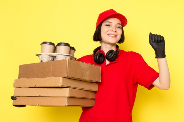 Une vue de face jeune courrier en uniforme rouge gants noirs et bonnet rouge tenant des boîtes de nourriture et une tasse de café