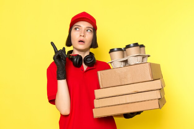 Une vue de face jeune courrier en uniforme rouge gants noirs et bonnet rouge tenant des boîtes de nourriture et des tasses à café