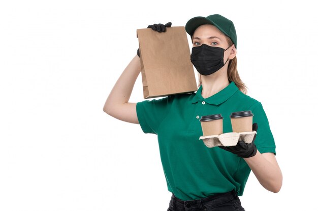 Une vue de face jeune courrier en uniforme vert gants noirs et masque noir tenant des tasses à café et des paquets