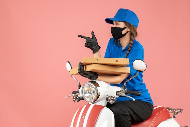 Vue de face d'une jeune coursière curieuse portant un masque médical et des gants assis sur un scooter livrant des commandes sur fond de pêche pastel