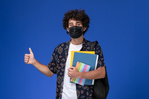 Vue de face jeune étudiant masculin portant un masque noir avec sac à dos contenant des fichiers et un cahier sur fond bleu.