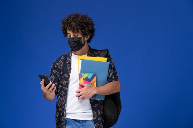 Vue de face jeune étudiant masculin portant un masque noir avec sac à dos contenant des fichiers et téléphone sur fond bleu