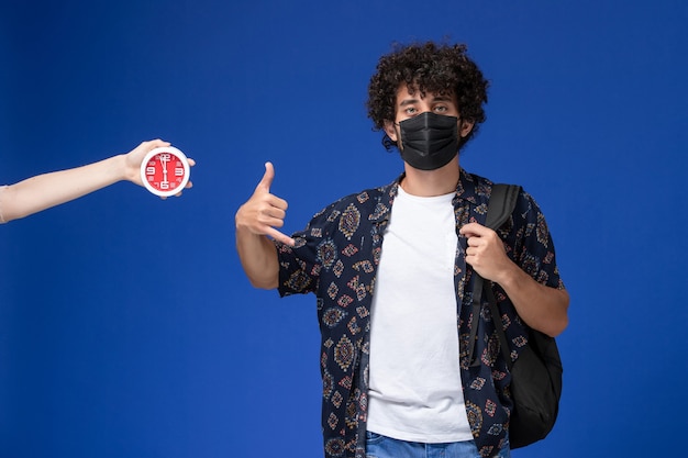 Vue de face jeune étudiant masculin portant un masque noir avec sac à dos posant sur fond bleu clair.