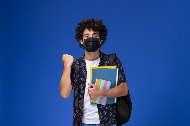 Vue de face jeune étudiant masculin portant un masque noir avec sac à dos tenant un cahier et des fichiers sur le fond bleu clair.