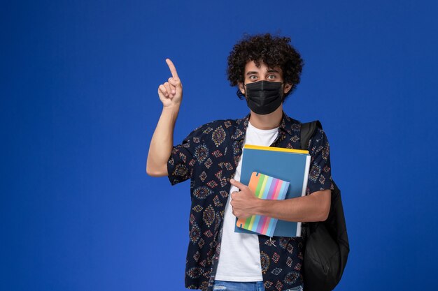 Vue de face jeune étudiant masculin portant un masque noir avec sac à dos tenant un cahier et des fichiers sur le fond bleu.