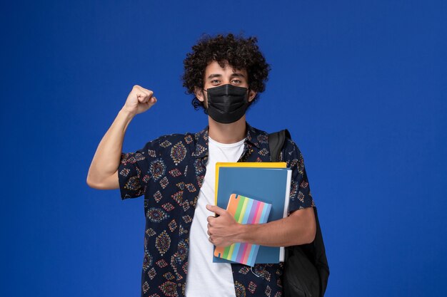 Vue de face jeune étudiant masculin portant un masque noir avec sac à dos tenant un cahier et des fichiers se réjouissant sur fond bleu.