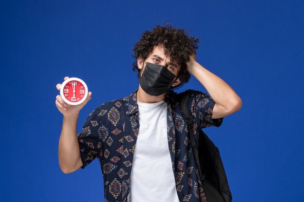 Vue de face jeune étudiant masculin portant un masque noir avec sac à dos tenant des horloges sur un bureau bleu.