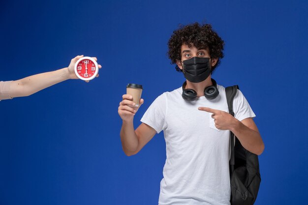 Vue de face jeune étudiant masculin en t-shirt blanc portant un masque noir et tenant du café sur le fond bleu.