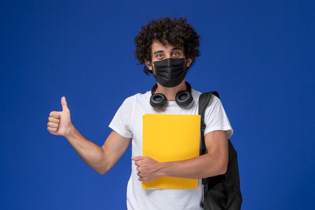 Vue de face jeune étudiant masculin en t-shirt blanc portant un masque noir et tenant des fichiers jaunes sur fond bleu clair.