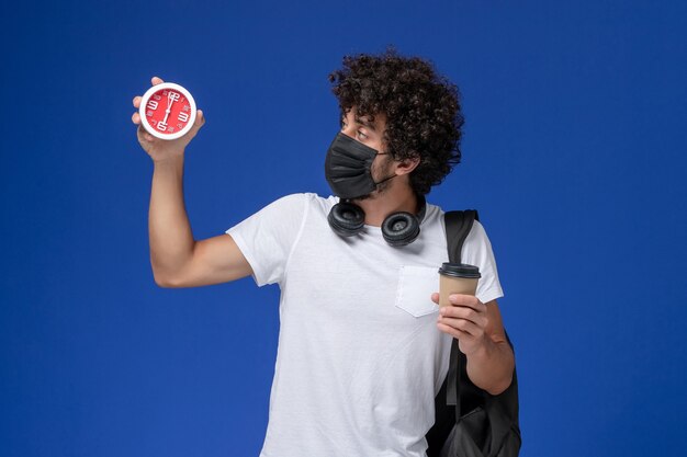 Vue de face jeune étudiant masculin en t-shirt blanc portant un masque noir et tenant une tasse de café avec horloge sur le fond bleu.