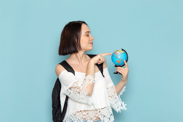 Vue de face jeune étudiante en chemise blanche jeans bleu et sac noir tenant petit globe rond sur l'espace bleu université étudiante