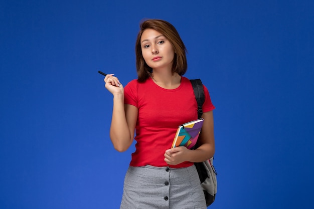 Vue de face jeune étudiante en chemise rouge portant un sac à dos tenant un cahier sur fond bleu clair.