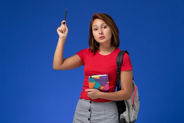 Vue de face jeune étudiante en chemise rouge portant un sac à dos tenant un cahier sur fond bleu clair.